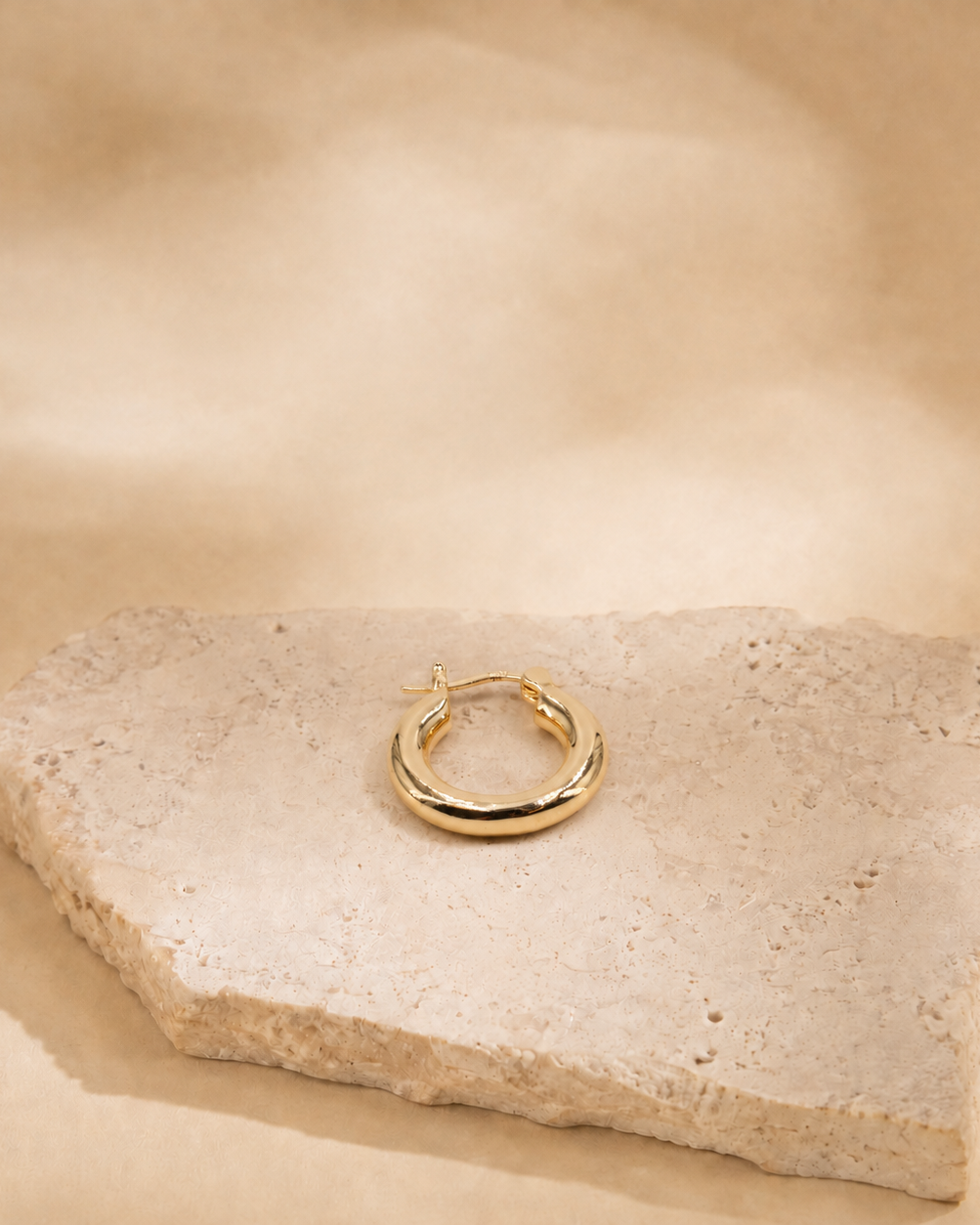Alba gold-plated hoop earring laid flat on a cream stone surface, showing its circular outline and clasp.