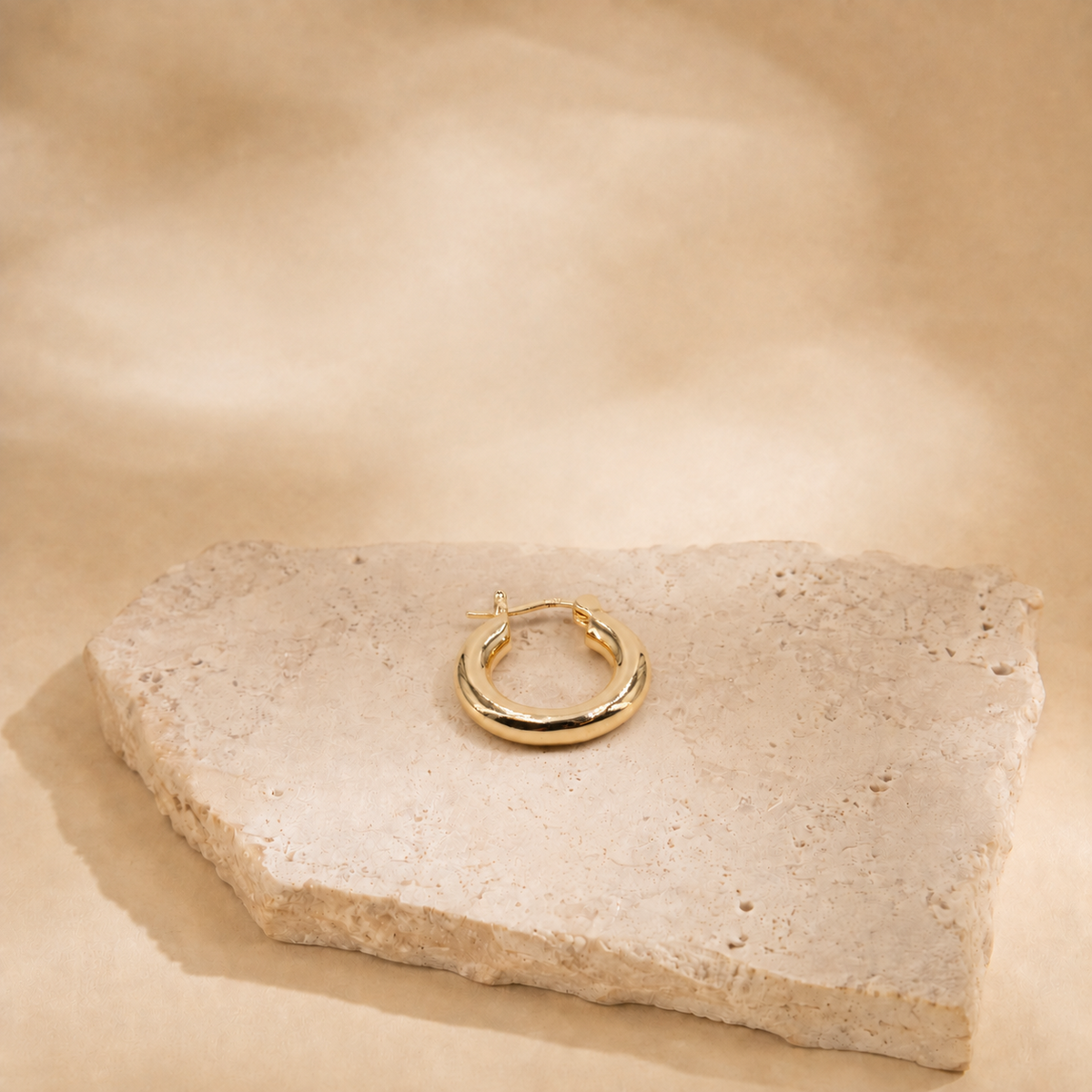 Alba gold-plated hoop earring laid flat on a cream stone surface, showing its circular outline and clasp.