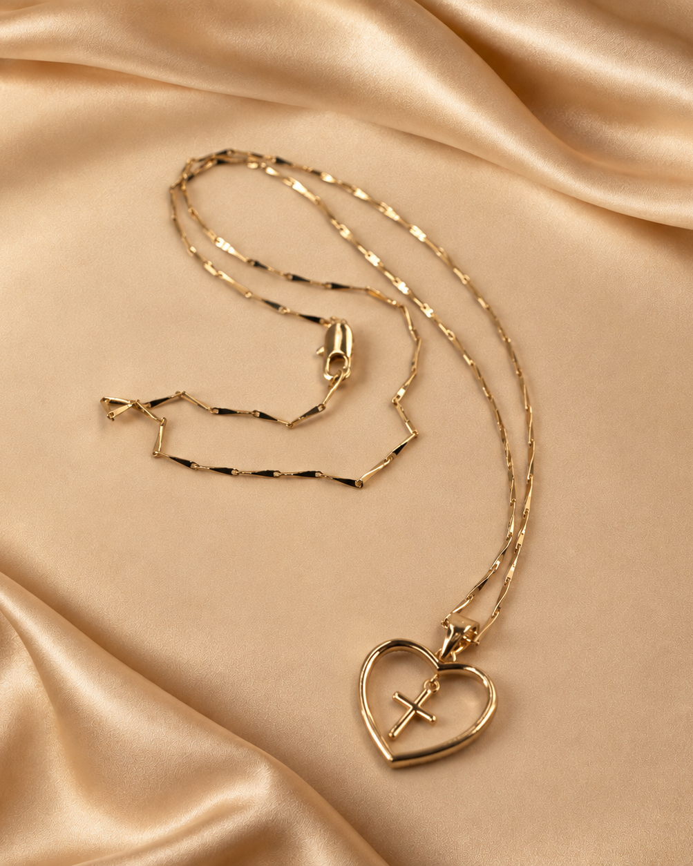 Amara 14K gold-plated necklace with an open-heart pendant and suspended cross, shown with its fine angular chain on champagne fabric.