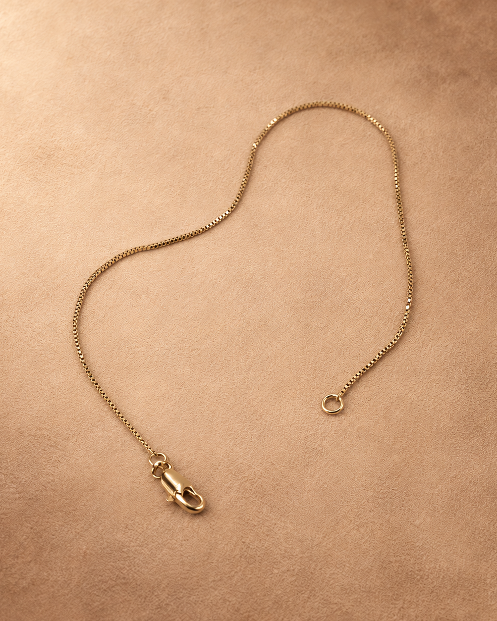 Linea fine chain bracelet laid open, with the lobster clasp and end ring separated on a warm textured background.