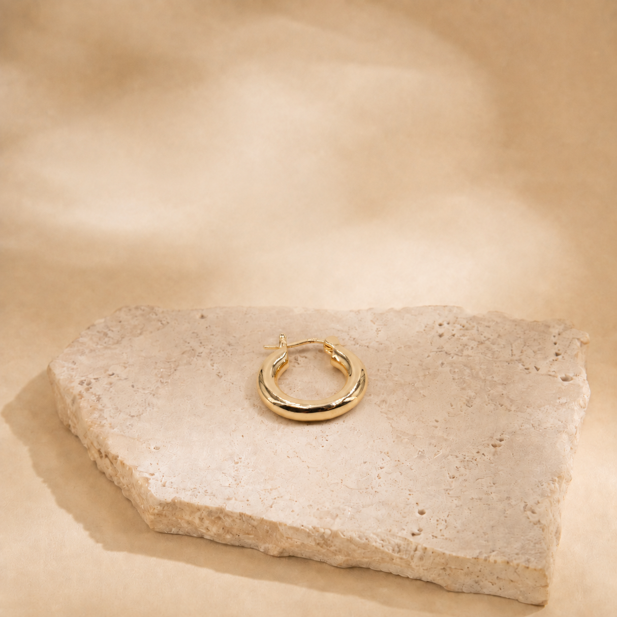 Alba gold-plated hoop earring laid flat on a cream stone surface, showing its circular outline and clasp.