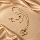 Amara 14K gold-plated necklace with an open-heart pendant and suspended cross, shown with its fine angular chain on champagne fabric.
