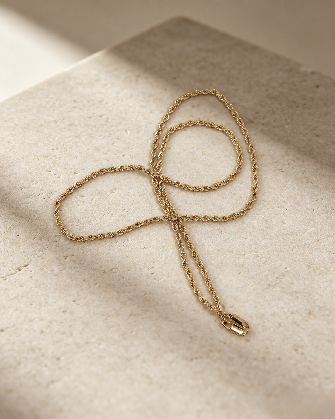 Corda rope chain necklace arranged in loose curves on a cream stone surface, with the polished clasp at the lower right.