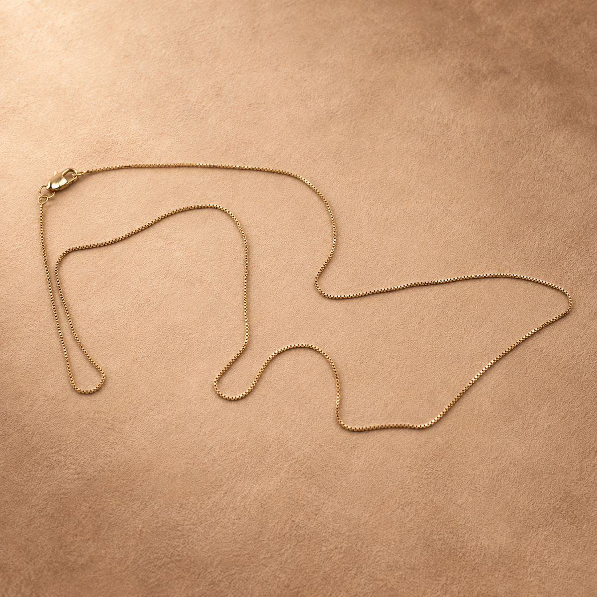 Linea box chain necklace arranged in loose curves on a warm textured surface, with the clasp at the upper left.